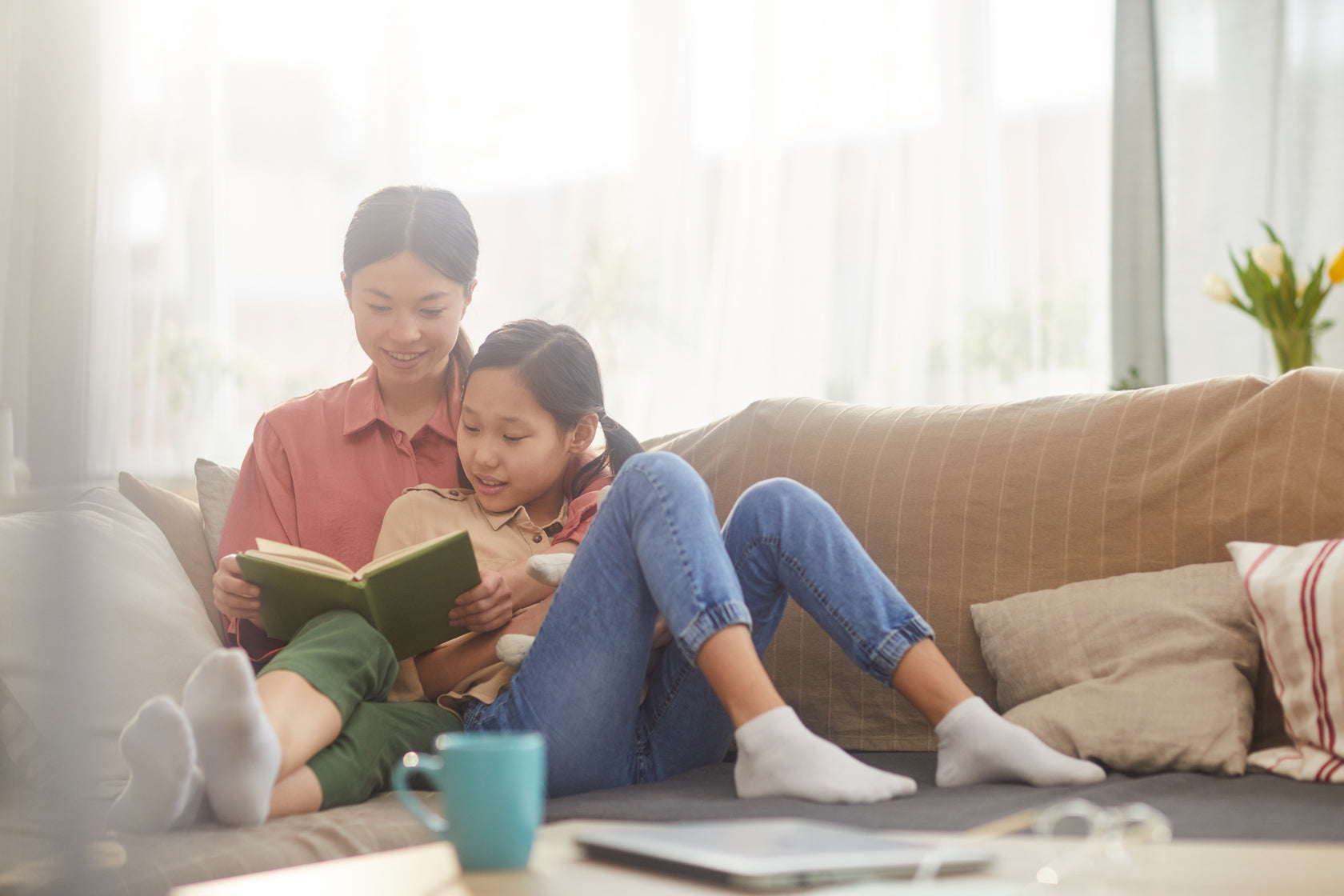 A mother and daughter sitting together on a sofa at home, sharing a book in soft natural light