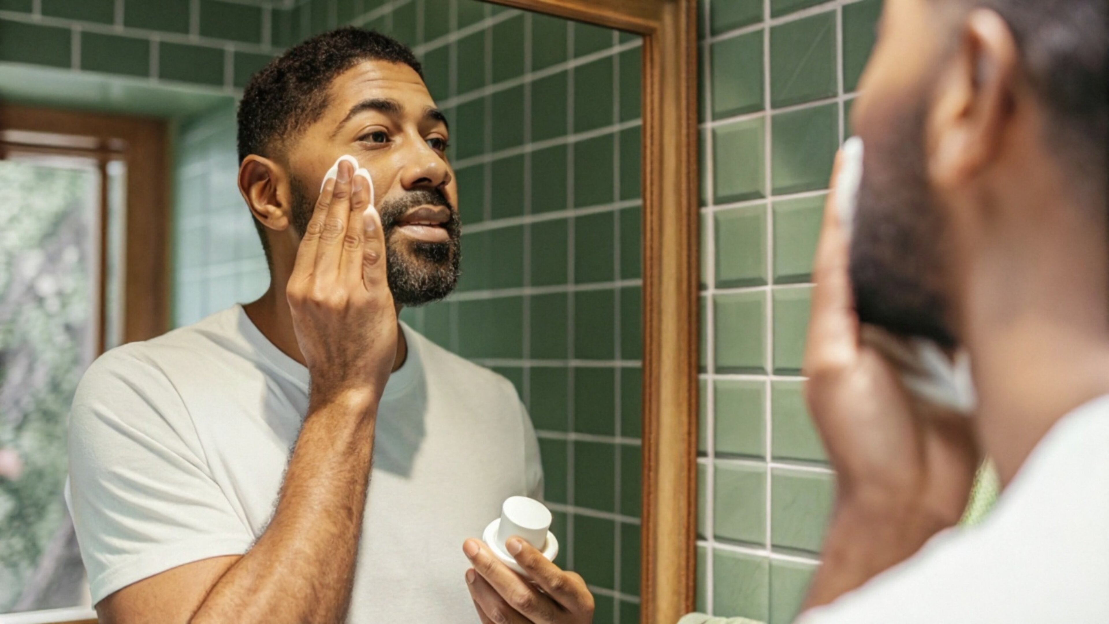 A man applying moisturiser to his face from a small jar while looking in a bathroom mirror, photographed in warm natural light.