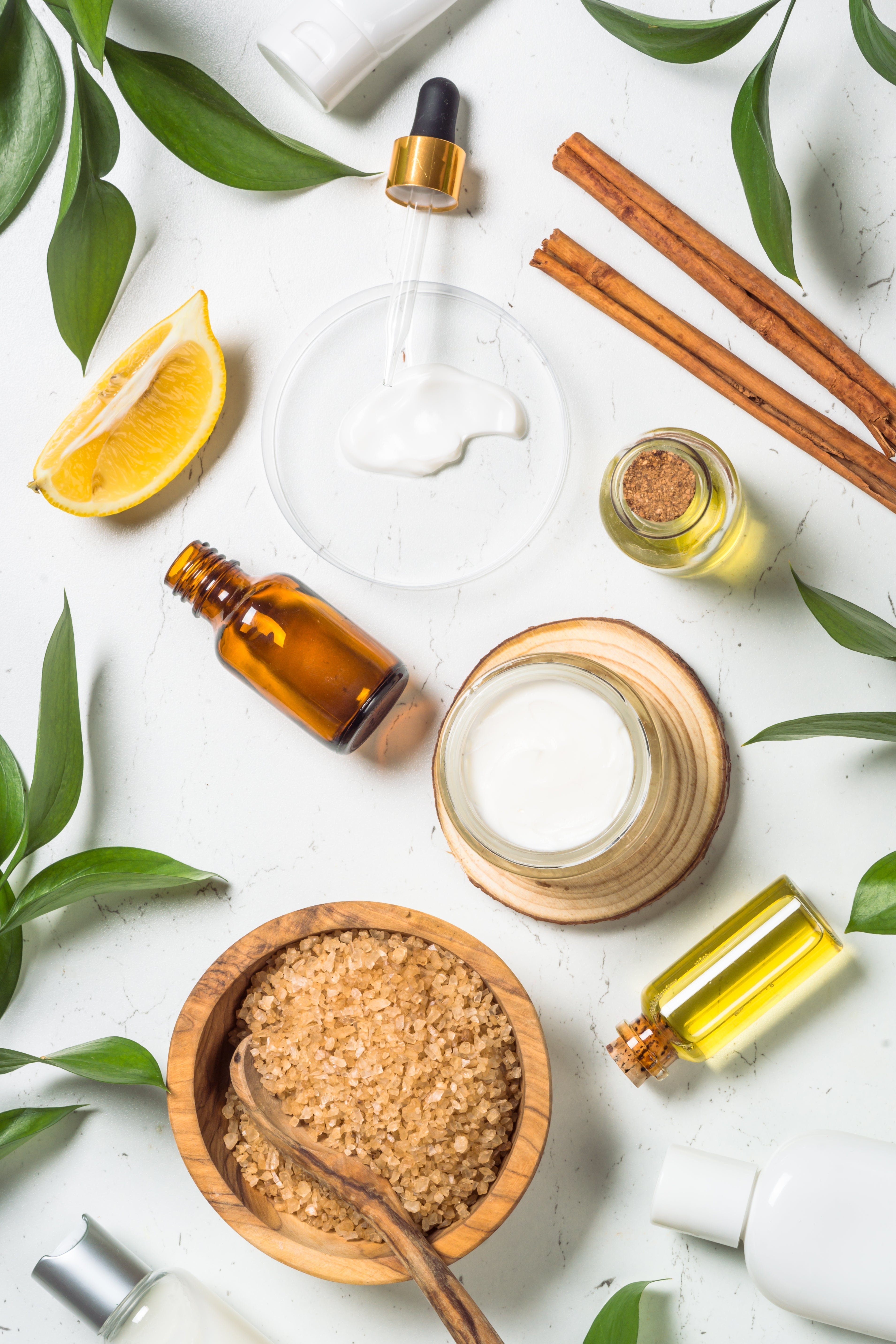 A flat-lay of natural skincare ingredients including amber oil bottles, a dropper, plant extracts, and a wooden bowl of brown sugar crystals, surrounded by green botanical leaves on a white surface.