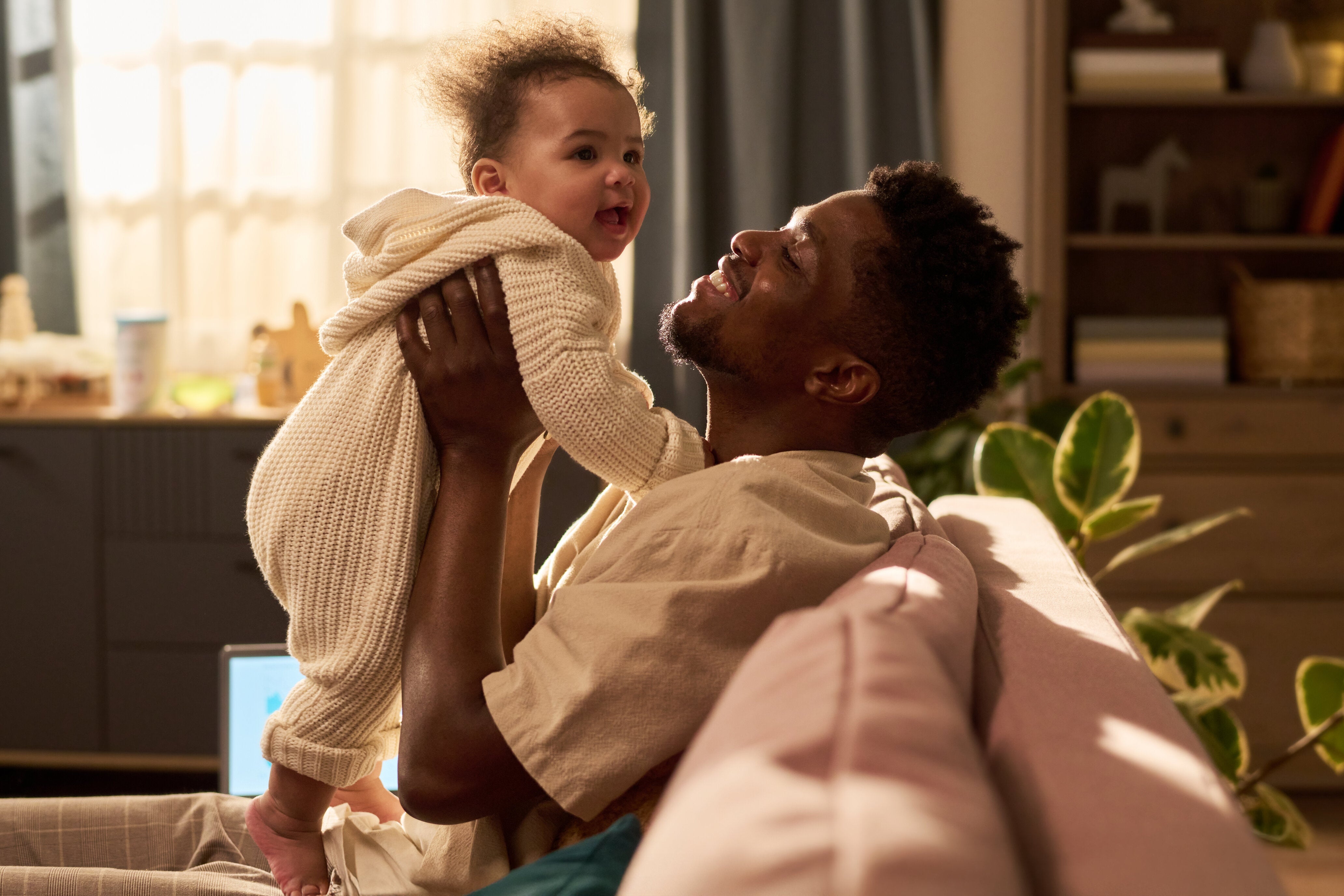 A father holding his baby up and smiling at each other, photographed in warm natural light at home.
